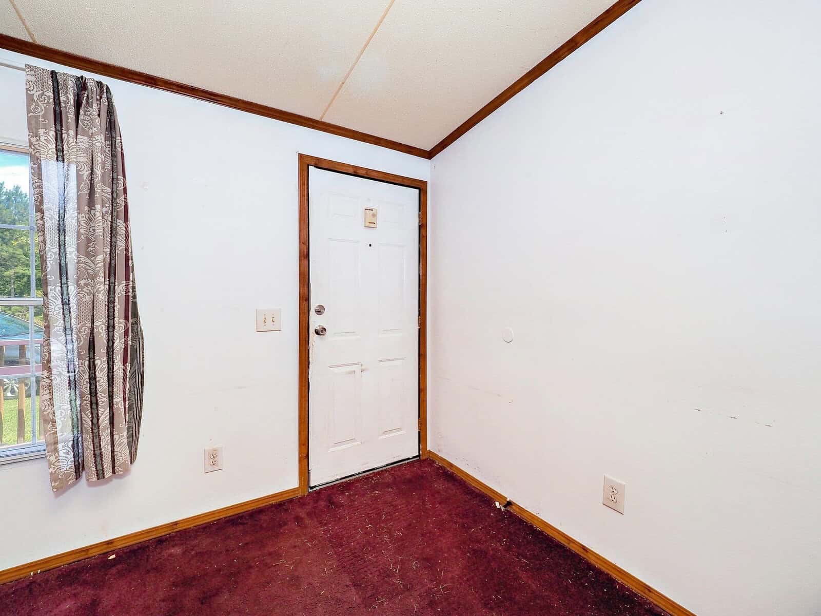 A small room with white walls and a maroon carpet, featuring a white door and a window with patterned curtains on the left. A light switch is on the wall near the door. The room appears minimally furnished.