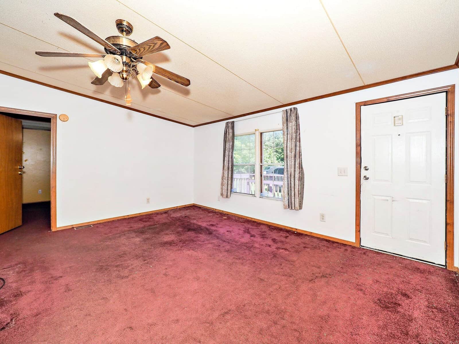 An empty room with purple carpet and white walls, featuring a ceiling fan with lights. There's a window with floral curtains and a white door on the right. An open doorway leads to another room. The room is well-lit by natural light.