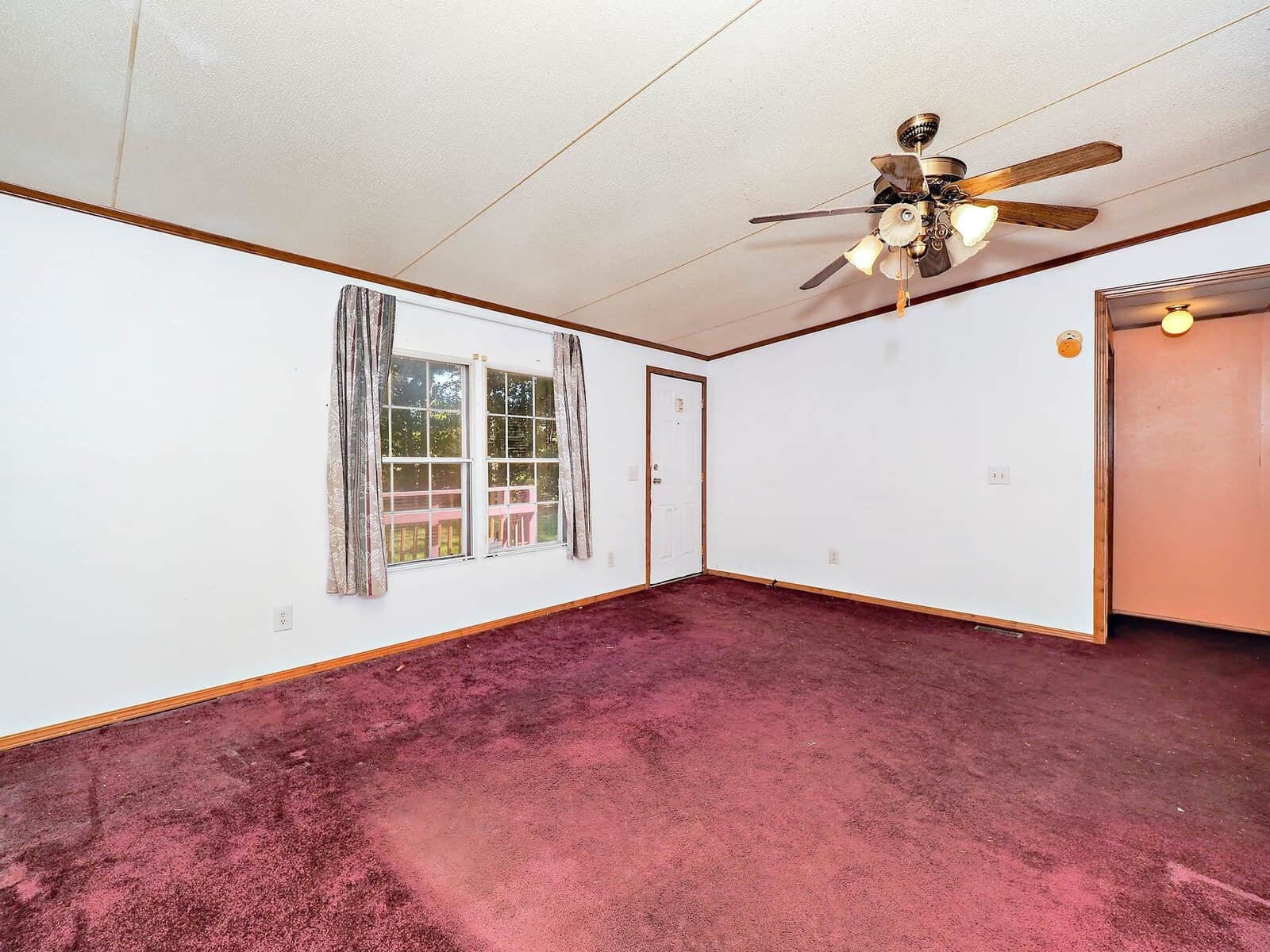 A spacious room with white walls and a maroon carpet. A large window and door are on one side, letting in natural light. A ceiling fan with lights is centered on the ceiling. A hallway is visible to the right.