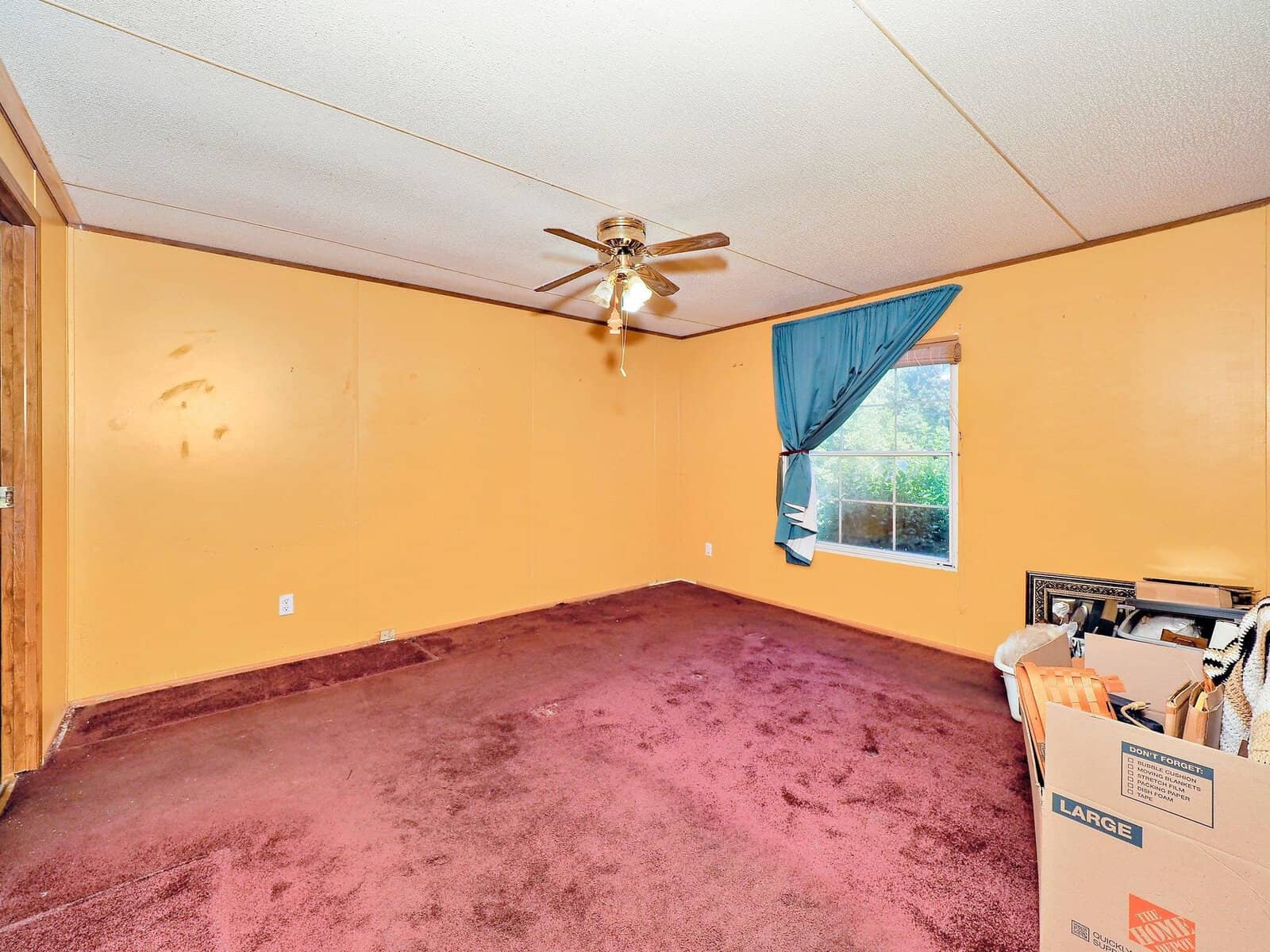 A room with mustard yellow walls and a mauve carpet. There is a ceiling fan and a window with blue curtains. A box filled with various items sits in the corner. The room appears empty and unfurnished.