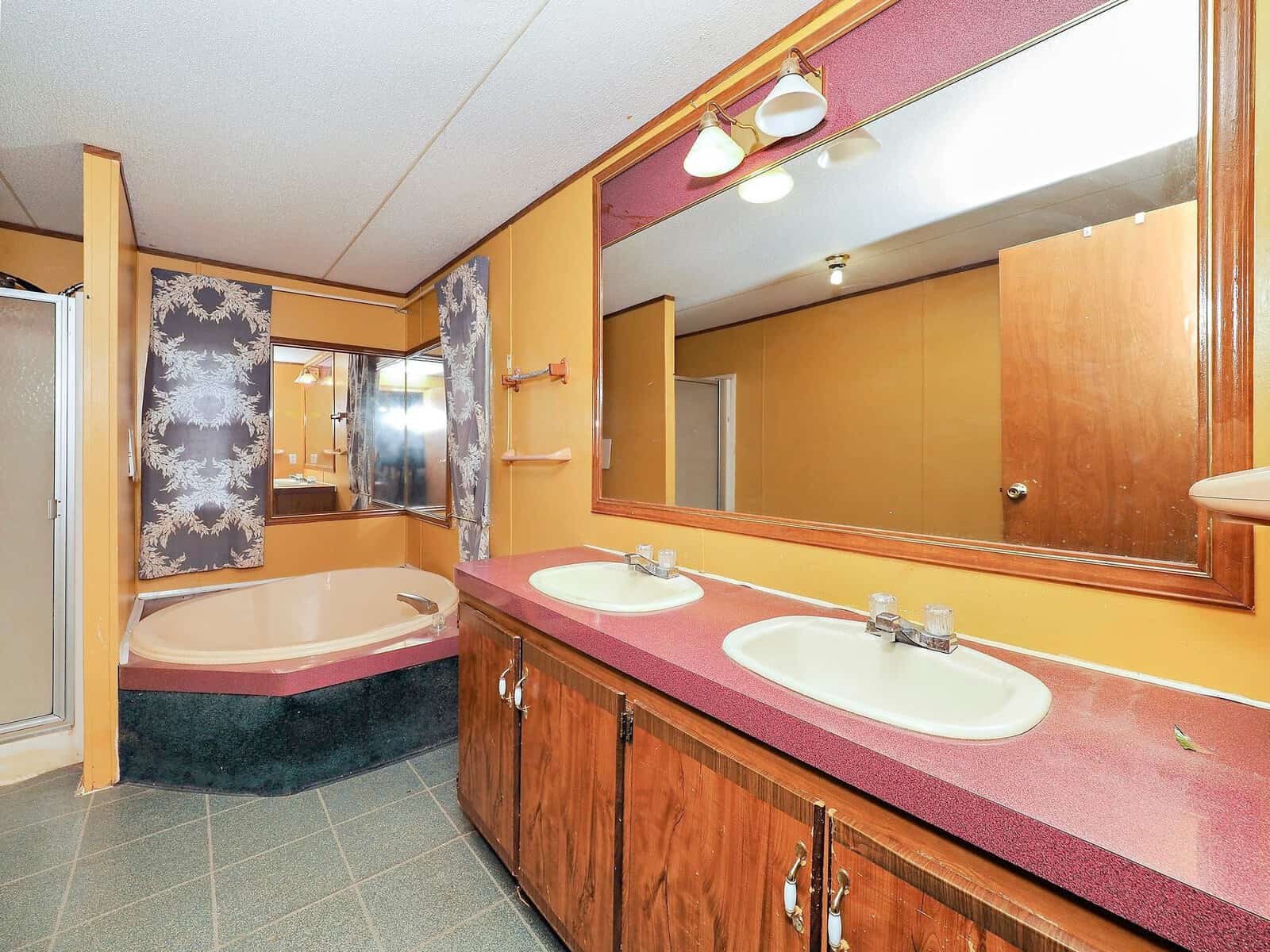 Bathroom with dual sinks, large mirror, pink countertop, and wooden cabinets. A corner bathtub with patterned curtains and a separate shower are also visible. The walls are yellow, and the floor is tiled.