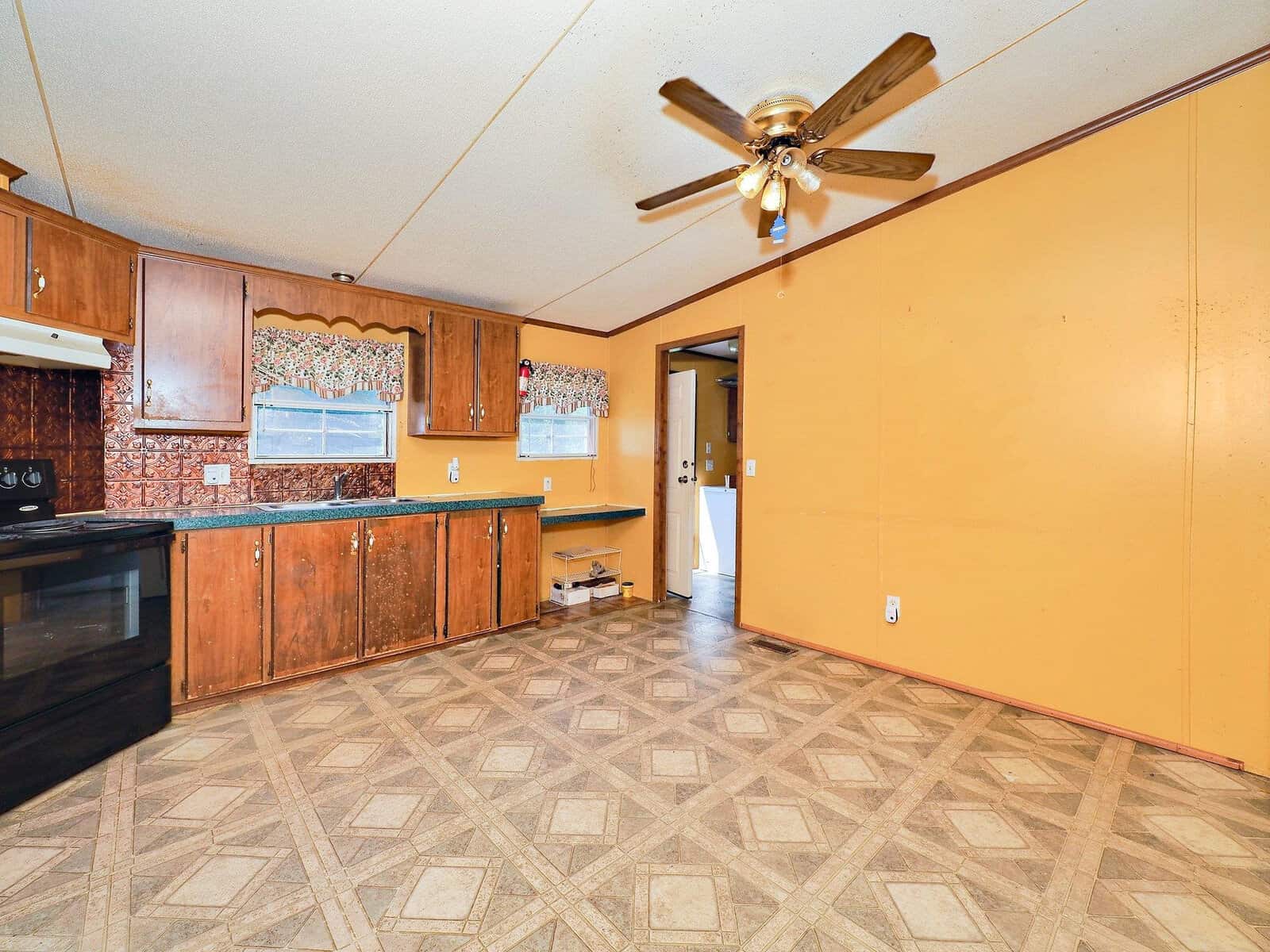 A kitchen with wood cabinets, a black stove, and a patterned tile floor. The walls are mustard-colored with a ceiling fan overhead. A doorway leads to another room with natural light illuminating the space.
