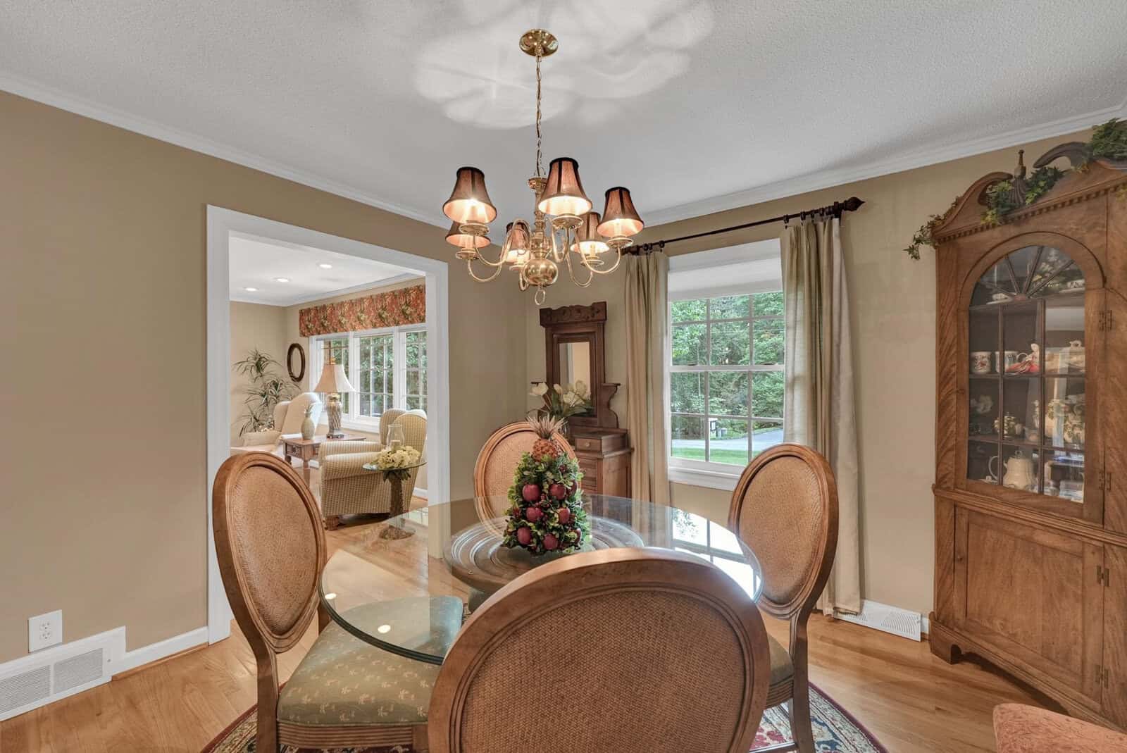 A well-appointed dining room with a round table, upholstered chairs, a wooden china cabinet, and a chandelier, featuring view to the adjacent room and outdoor through a window.