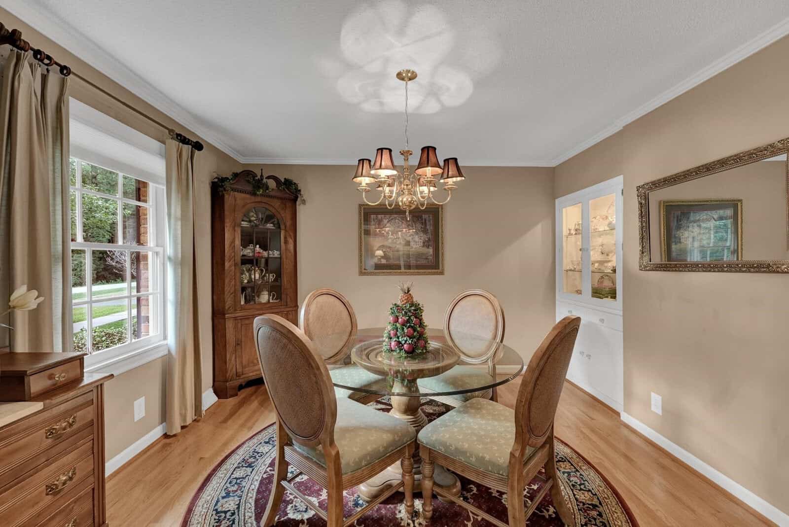 Traditional dining room with a glass-top table, four upholstered chairs, and a chandelier, featuring a corner cabinet, oriental rug, and views to the outdoors through windows.