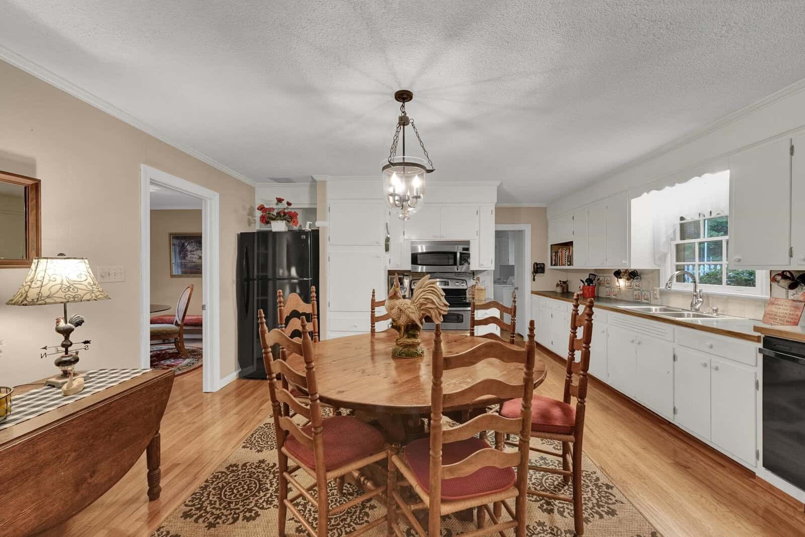 A well-lit dining room with a wooden table and chairs, leading into a kitchen with white cabinetry.