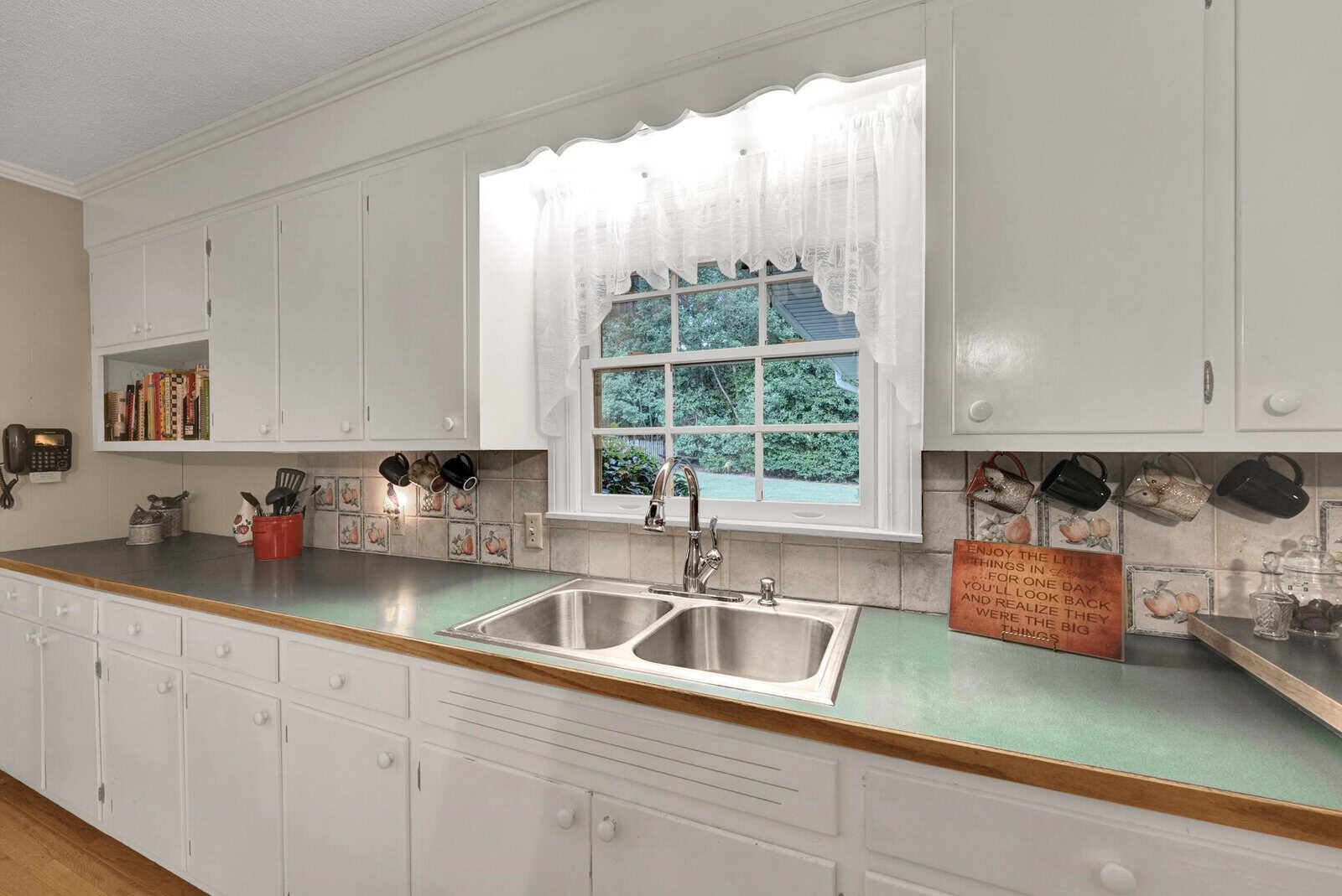 A well-lit kitchen with white cabinetry, stainless steel sink, and a window overlooking a yard.