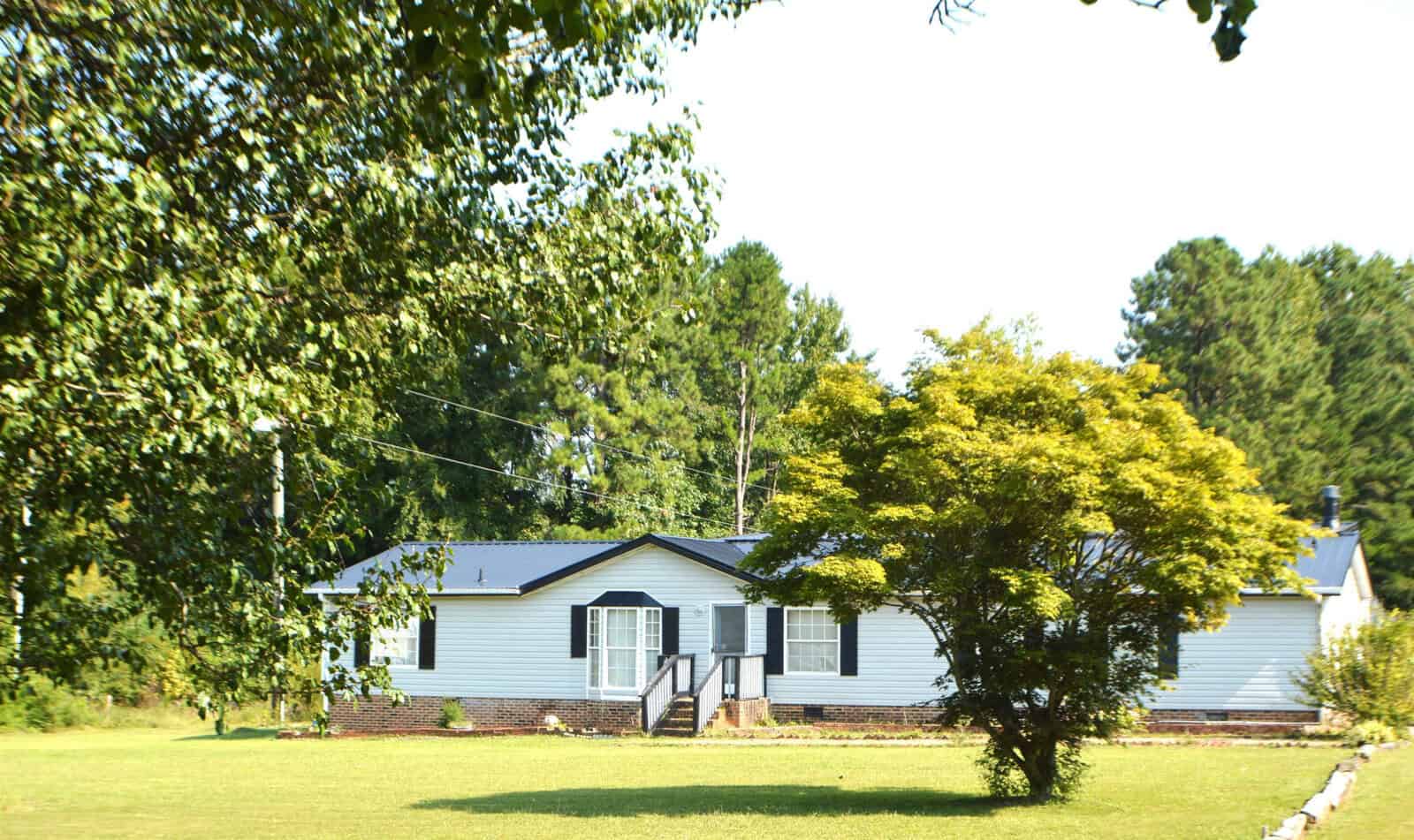 Single-story blue house with white trim, featuring a small porch, surrounded by greenery and a clear sky.