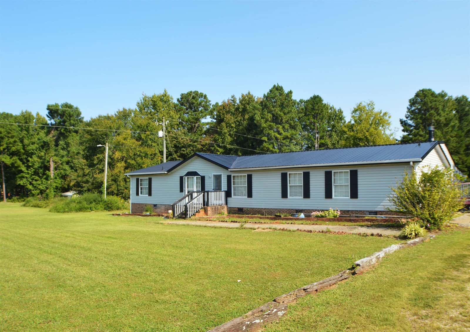 A single-story blue house with white trim and a dark roof, featuring a small porch with stairs, set in a large, grassy lawn with surrounding trees under a clear blue sky.