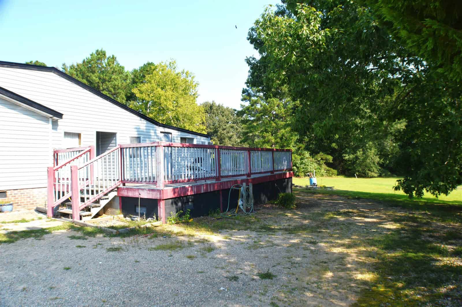 A single-story building with light siding features a red deck with white railing and a set of stairs; a large tree casts shade over the area and a lawn stretches into the background on a sunny day.