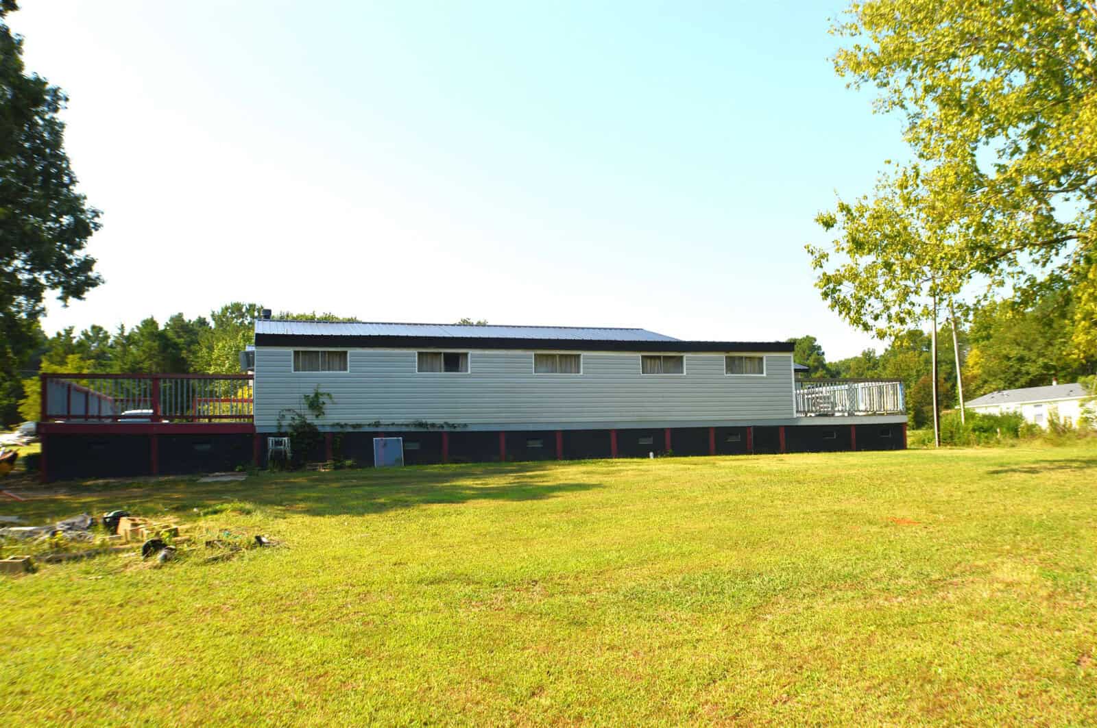 A two-story building with a gray exterior and red trim, featuring an expansive balcony and a fenced deck area, situated in a grassy field with trees in the background under a clear blue sky.