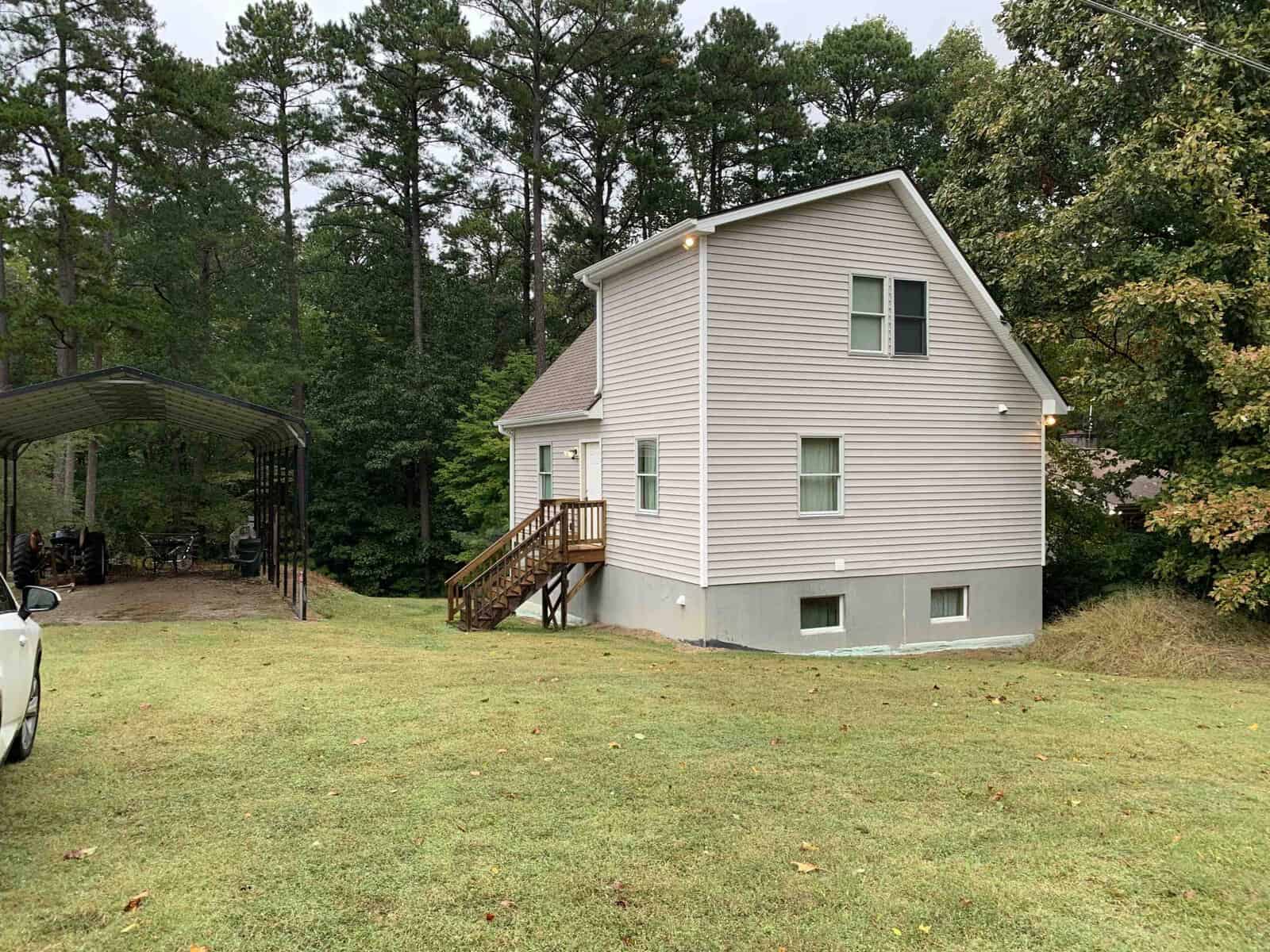 A two-story grey house with green shutters and a wooden deck, situated on a lawn with surrounding trees at 930 Cedar Cove Road, Henderson NC; to the left is a metal carport with