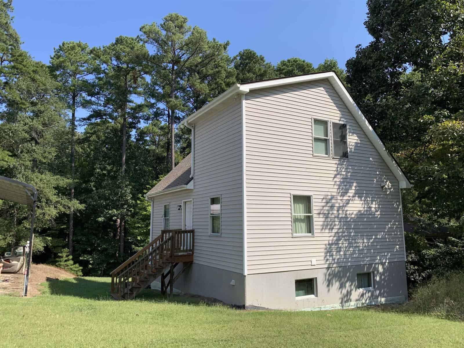 A two-story house with gray siding and white trim features a set of wooden stairs leading to the upper level, surrounded by lush green trees under a clear blue sky.