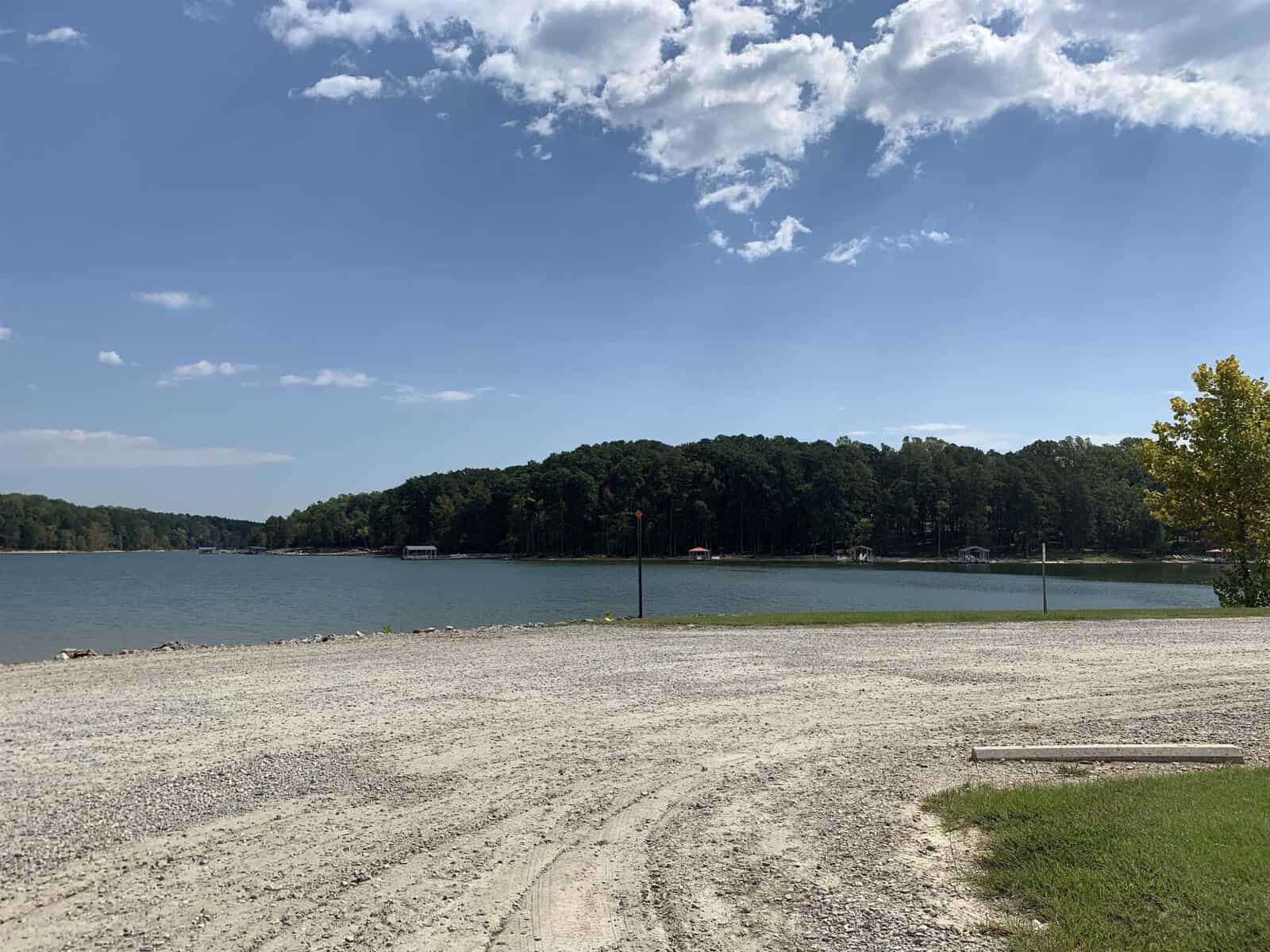 A tranquil lakeside view with a gravel shore in the foreground, surrounded by lush green trees under a partly cloudy sky.