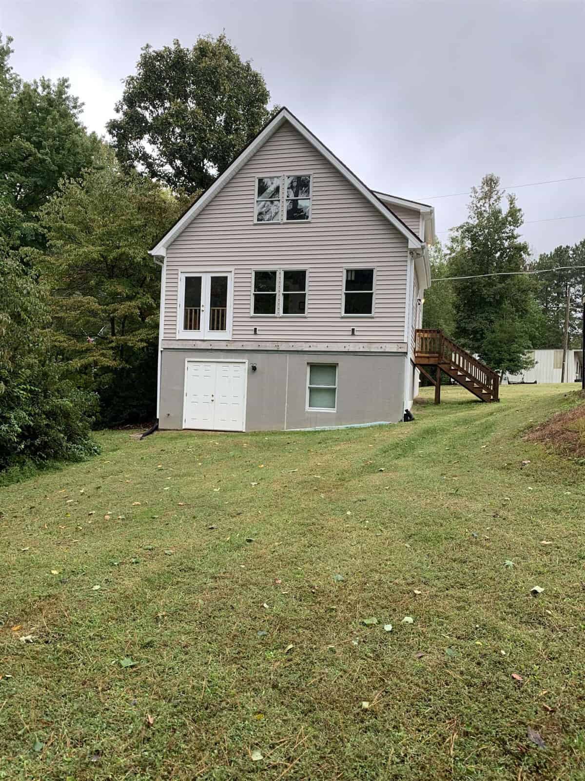 Two-story house with light siding and contrasting dark shutters, featuring a ground-level door and an upper deck with wooden stairs, situated on a grassy lawn with trees in the background, on a cloudy day.