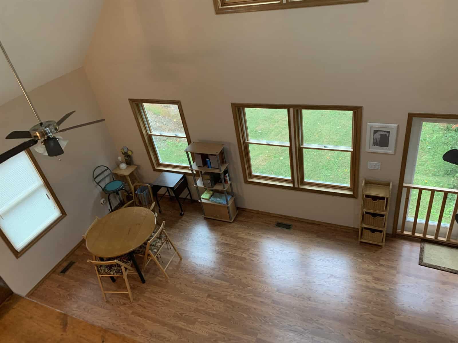 An overhead view of a brightly lit room with a high ceiling, featuring multiple windows, a ceiling fan, and minimal furniture including a table, chairs, and shelves.