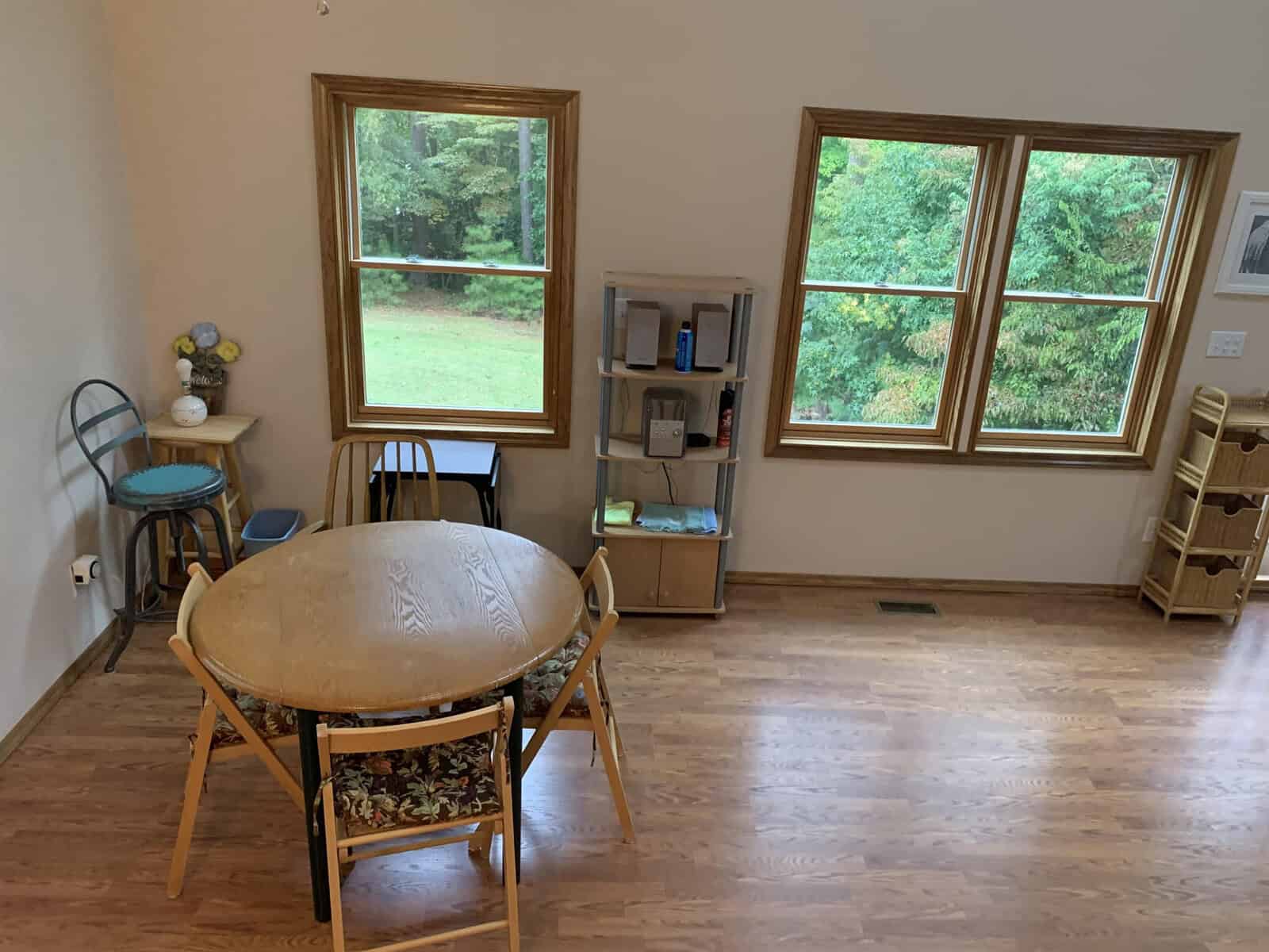 A simply furnished room with a circular table and chair, a bookshelf with items, a cat tree, and three windows showing a view of trees and a lawn.