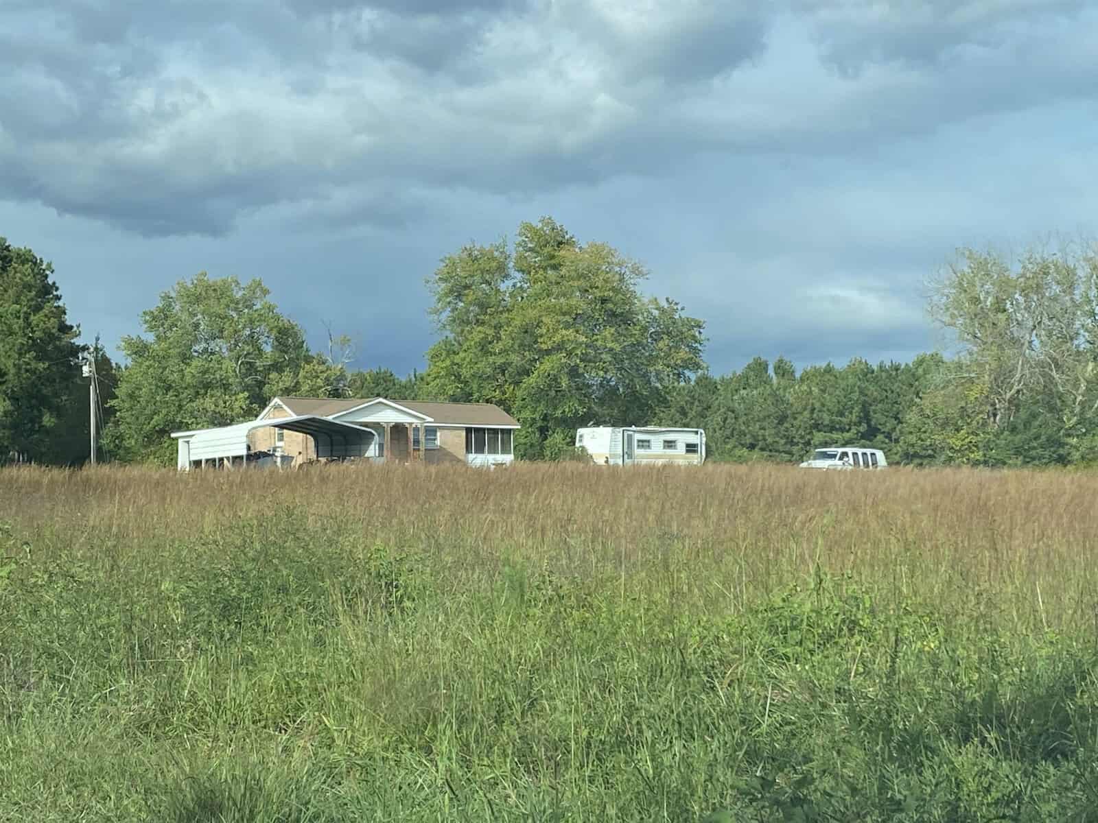 A rural residence with a blue roof is partially obscured by a field of tall grass, with a recreational vehicle and a white van parked to the right. clouds gather in the sky above, suggesting a possibility of rain.