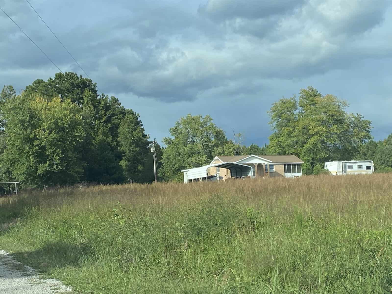 A rural landscape featuring a house and a mobile home amid tall grass under a cloudy sky.