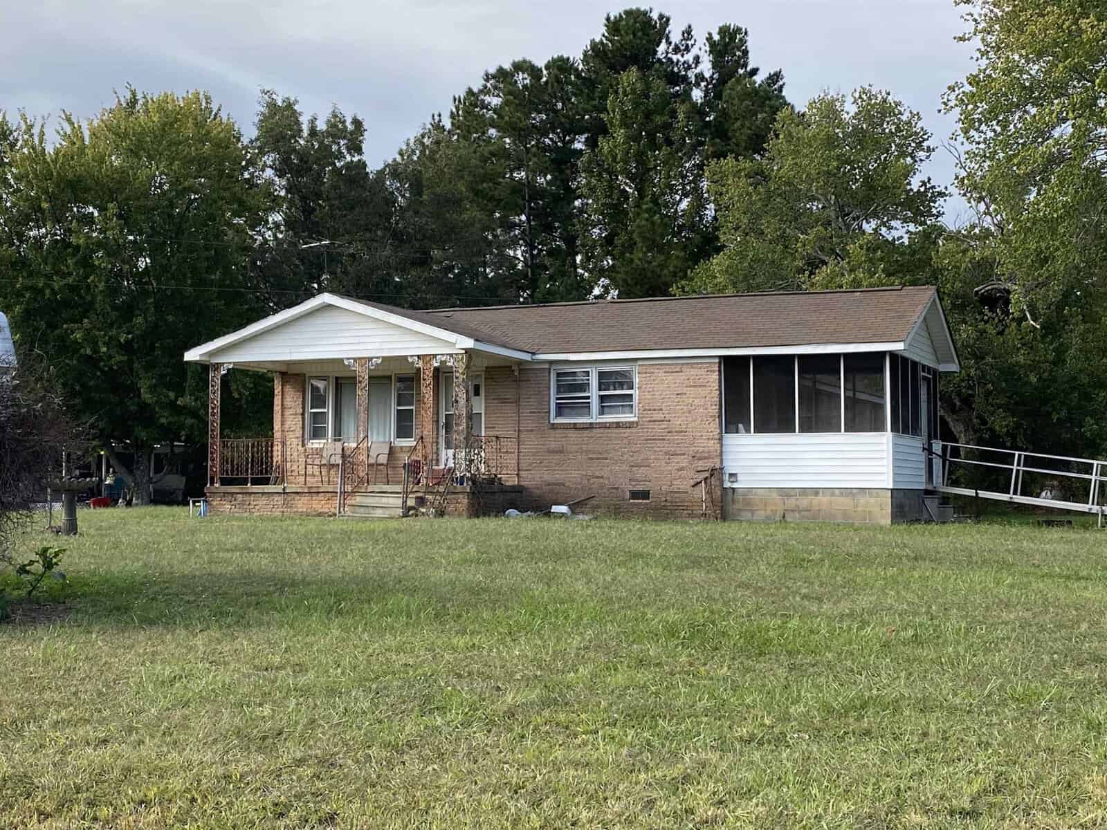 A single-story brick and siding house with a covered front porch and a wheelchair-accessible ramp, surrounded by a grassy yard with trees in the background.