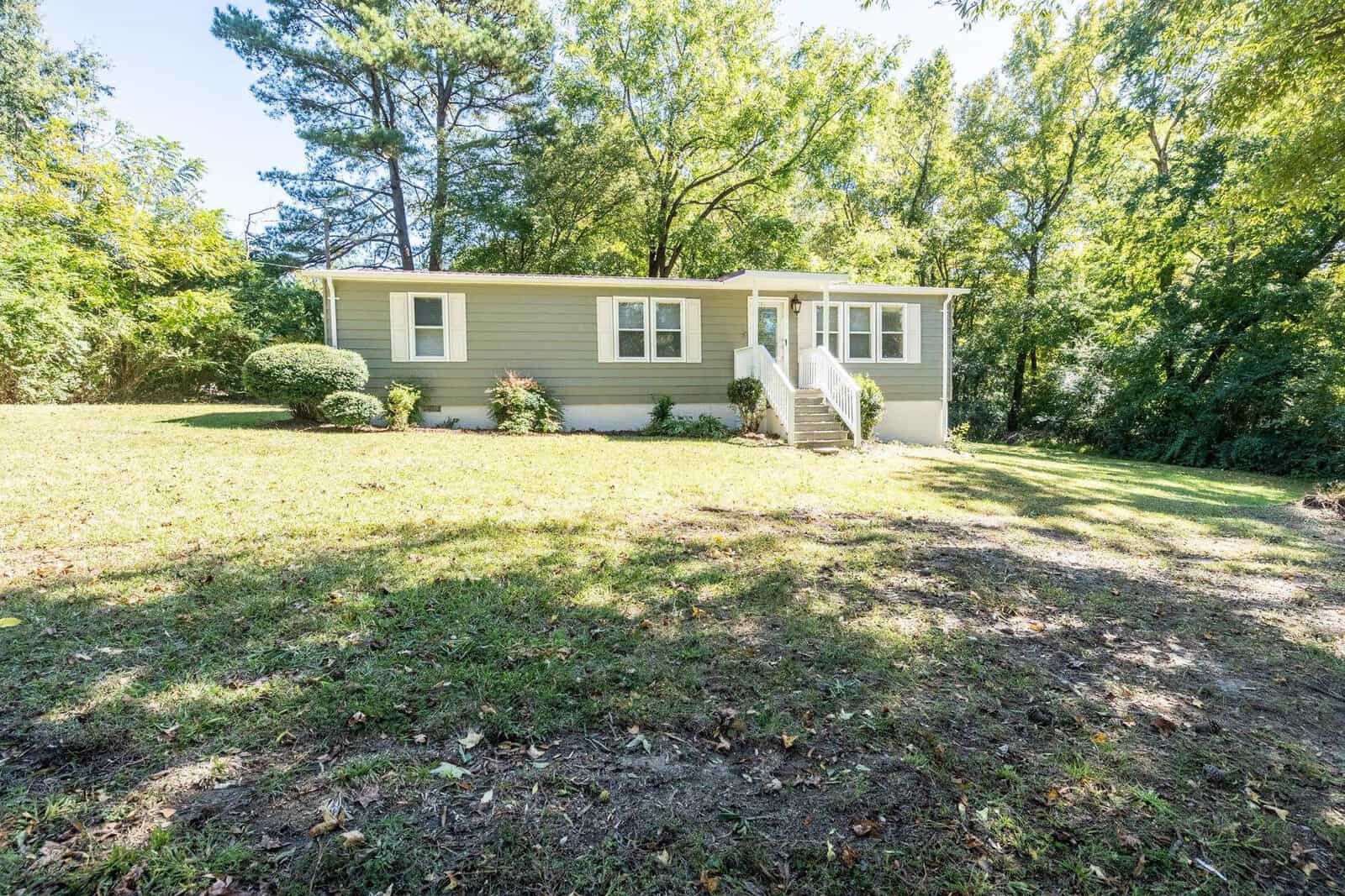 A single-story house with gray siding is positioned among trees on 109 Old Watkins Road, Henderson NC, featuring white stairs leading to a side entrance, on a sunny day with shadows cast across the lawn