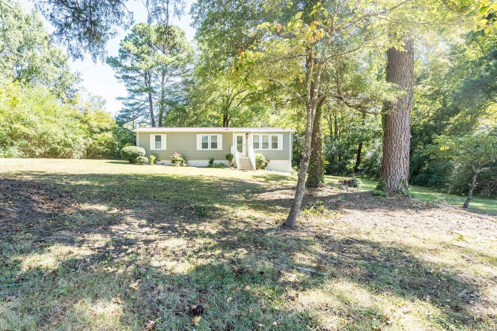A single-story green house with white trim, surrounded by trees and situated on a grassy lawn with a slightly sloping terrain, under a clear sky.