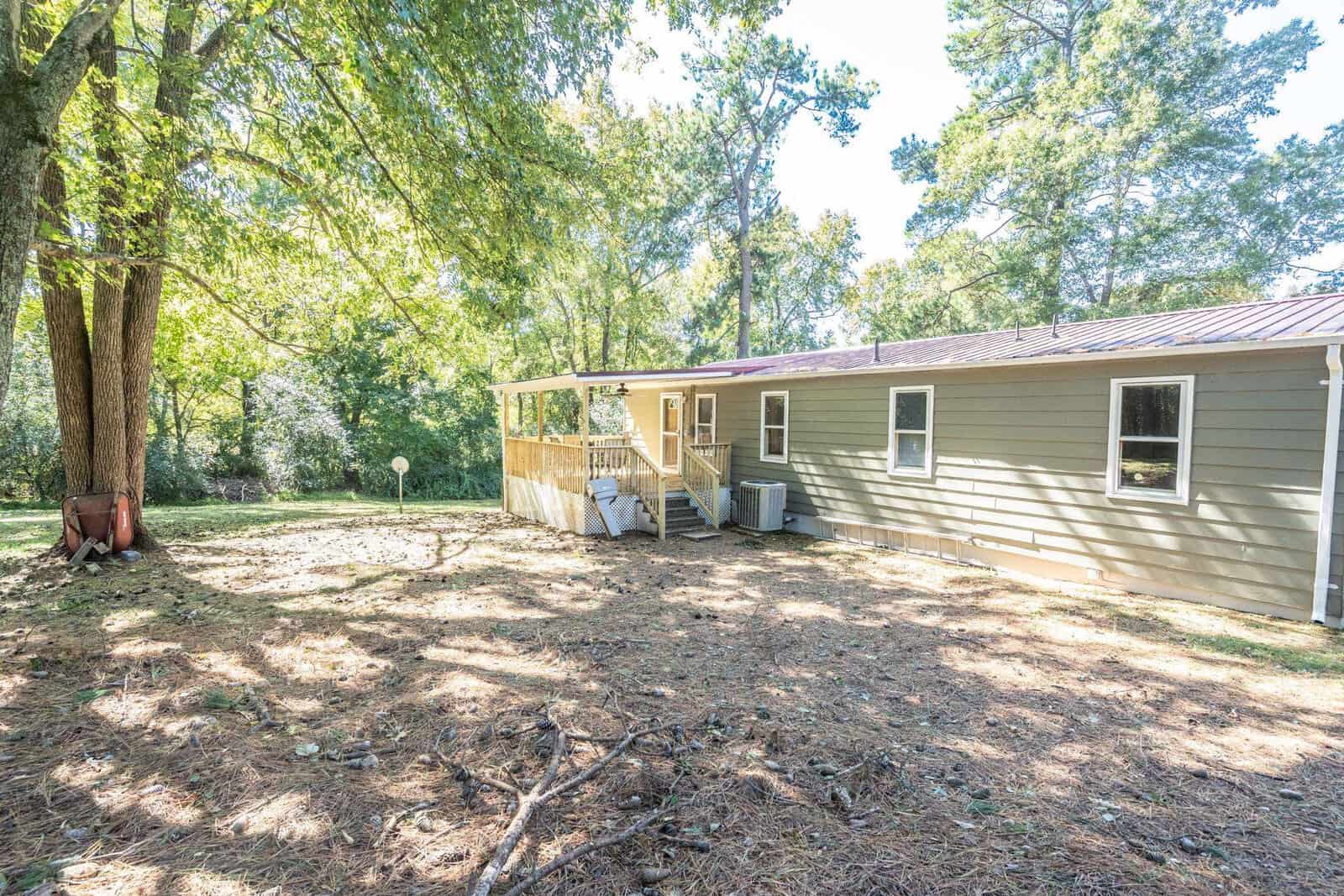 A single-story house with a wooden deck surrounded by trees and a clear yard area.