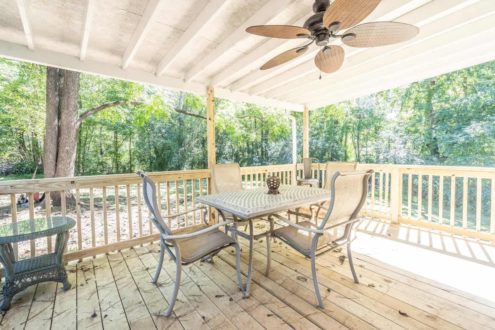 A spacious, covered outdoor deck with a table and four chairs set, surrounded by lush greenery, featuring ceiling fans above.