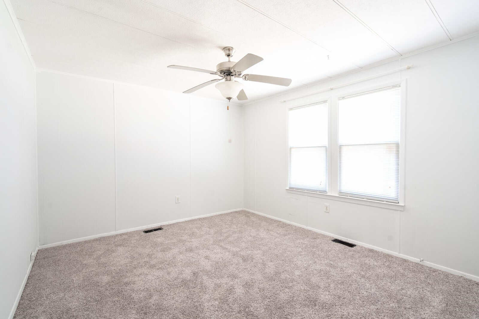 A sparsely furnished room with white walls, beige carpet, a ceiling fan, and two windows allowing in natural light.