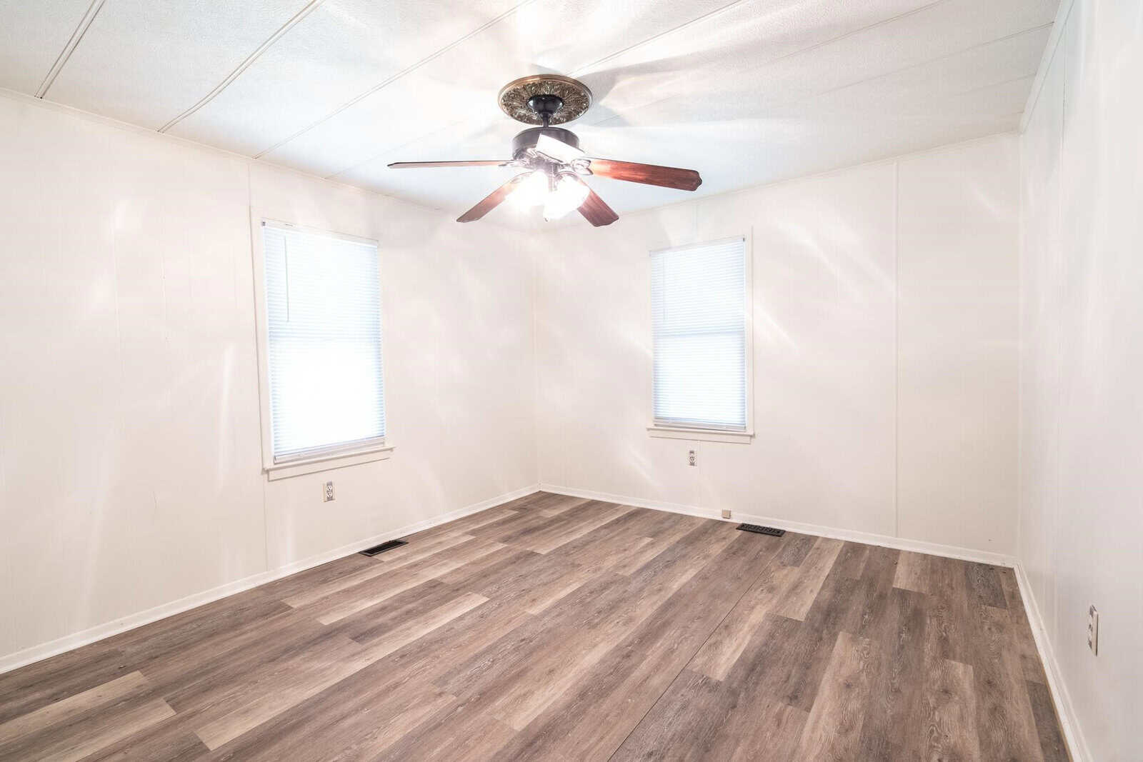 An empty room with light walls and dark wood laminate flooring featuring a ceiling fan with lights and two windows with blinds.