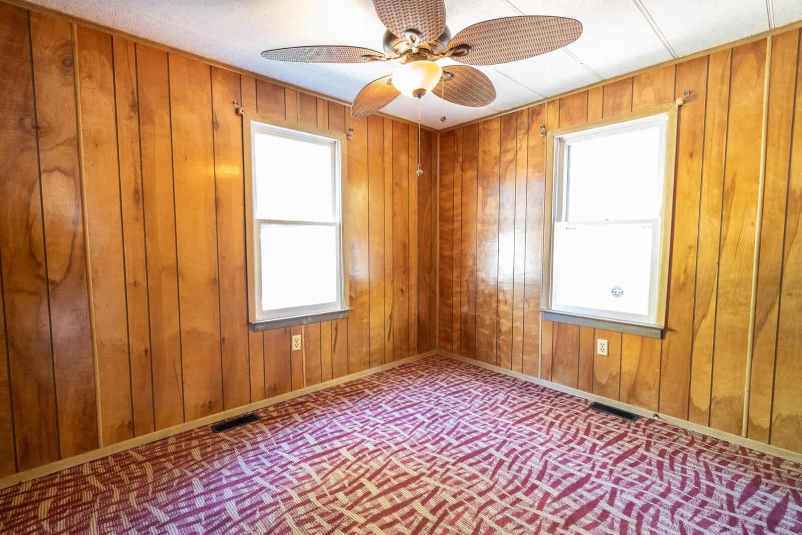 Empty room with wood paneling on walls, two windows, and a patterned carpet. a ceiling fan is mounted above.