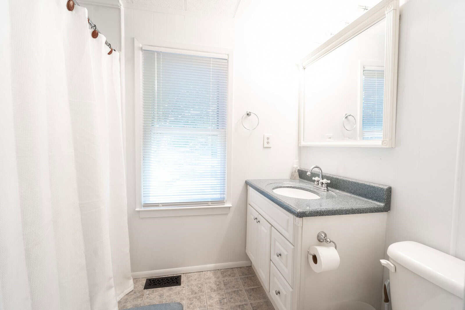 A clean and bright bathroom with a shower curtain, a window with blinds, a sink with a granite countertop, and a toilet.