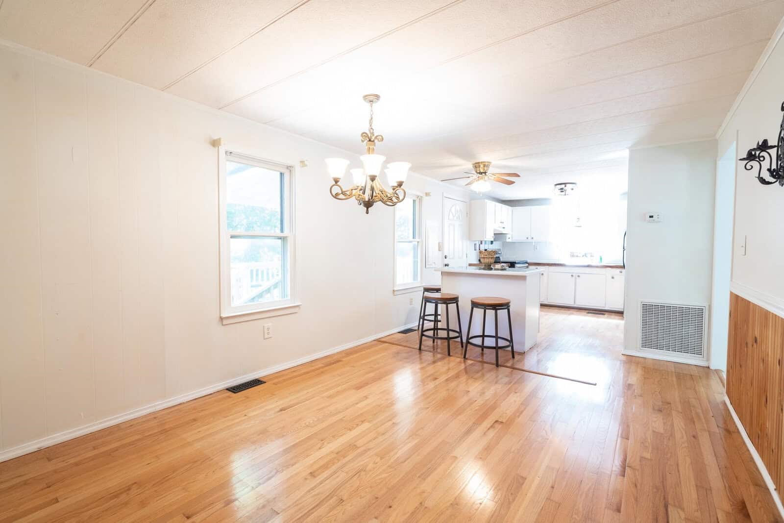 Spacious, well-lit room with hardwood floors, featuring a chandelier and an open kitchen with a bar seating area.