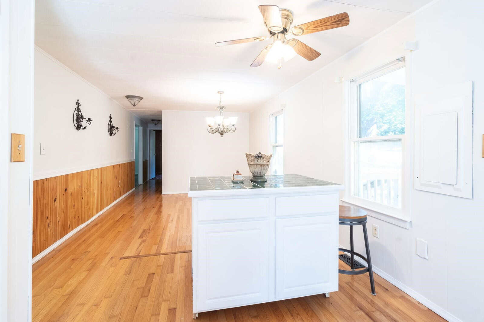 A bright, empty kitchen with wooden floors, white cabinetry, and a central island under a ceiling fan.