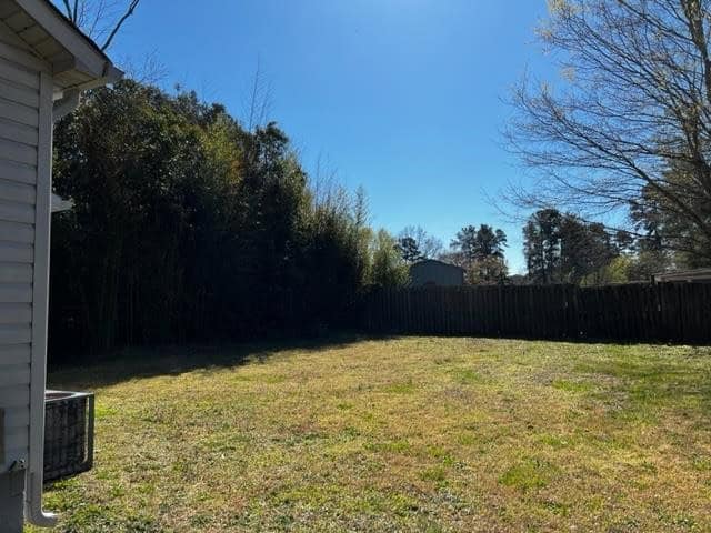 A sunny backyard with a trimmed lawn, bordered by a wooden fence, with a view of trees and a building in the distance. an air conditioning unit is visible to the left, adjacent to a house.