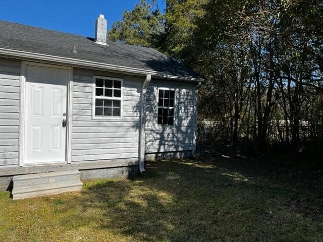 A small, single-story house with gray siding and a white door, featuring a small concrete step at the entrance, surrounded by grass and trees under a clear sky.