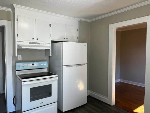 A compact kitchen with white appliances, including a refrigerator and an electric range, and white cabinetry with nickel handles. the walls are painted in a light gray color, and the kitchen opens to another room with hardwood flooring.