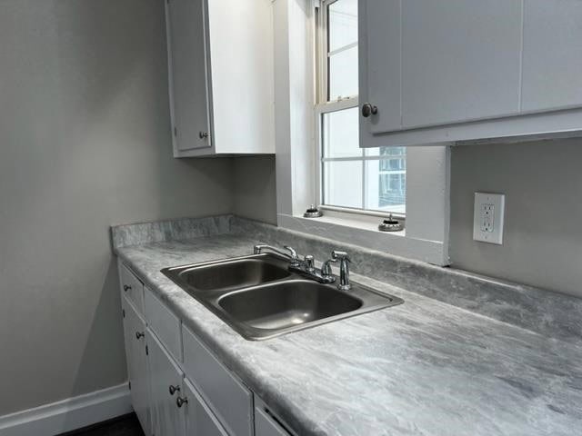 A corner of a kitchen with white cabinetry, gray countertop, and a stainless steel sink beneath a window.