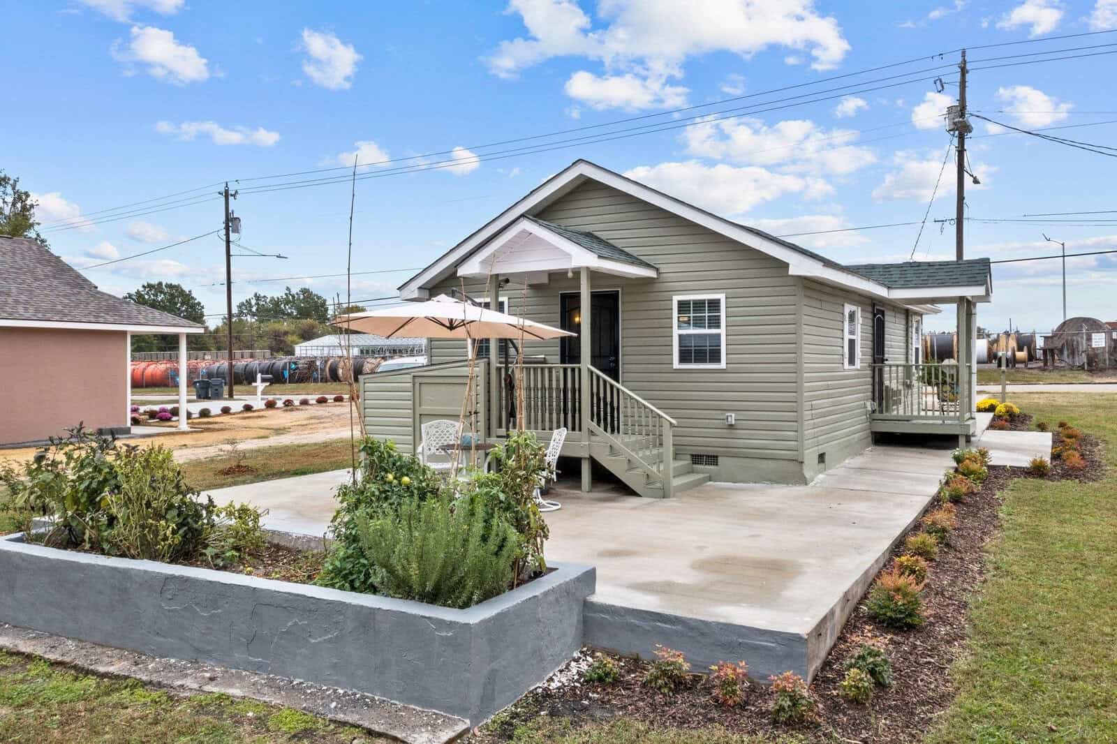 A single-story green house with a white front door and stairs leading up to a small covered porch. there is a concrete walkway in front and a raised garden bed with shrubs on the left. the house is situated in a neighborhood with clear skies above.