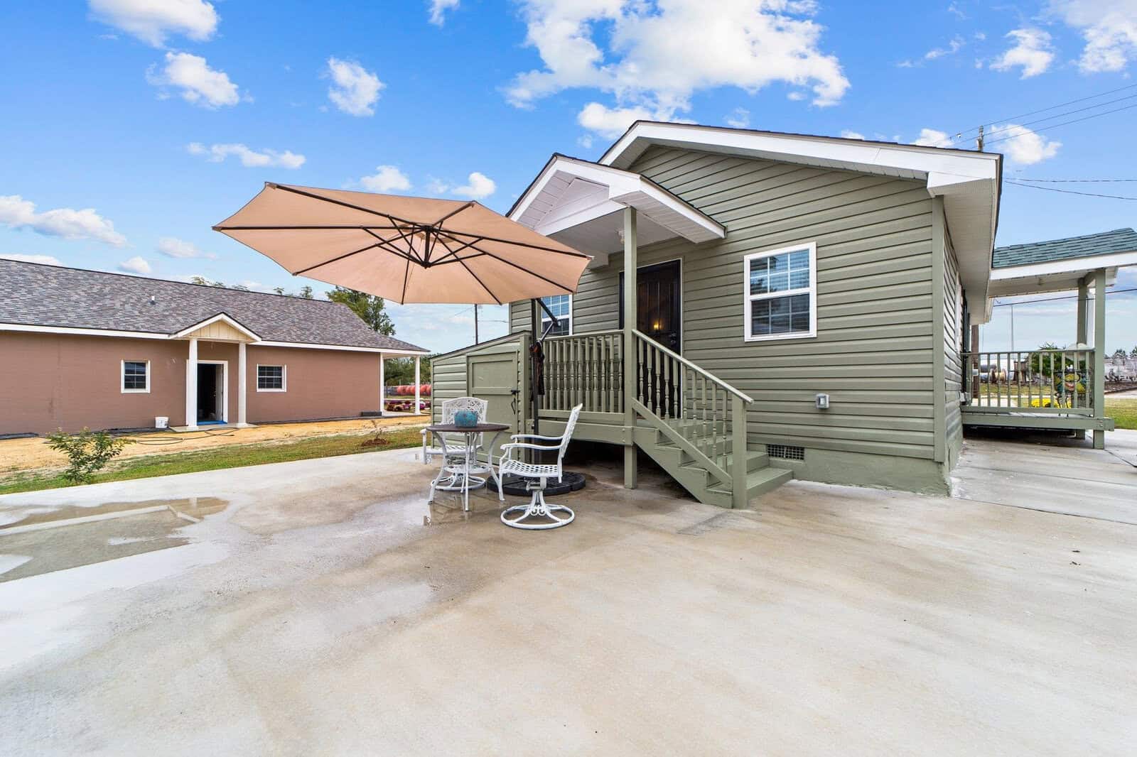 A small green painted house with white trim featuring a set of wooden steps leading to the front door, accompanied by a concrete patio area with a large umbrella and outdoor chairs.