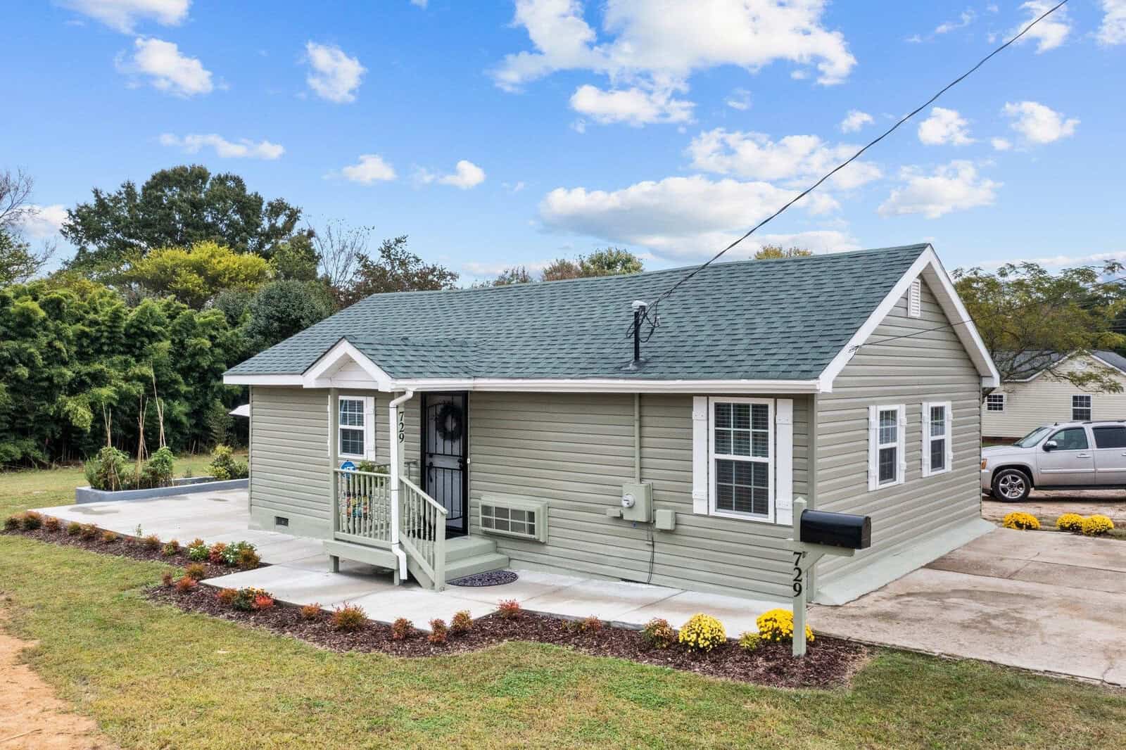 A small single-story gray house with a front porch and a shingled roof, featuring a neatly maintained front yard with flowerbeds and a concrete driveway with a parked car.