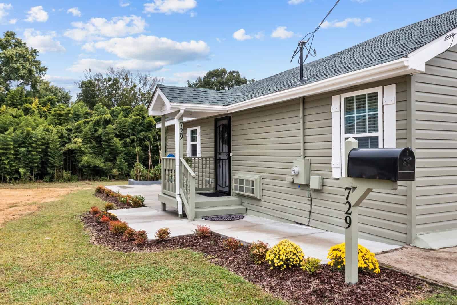 A single-story beige house with white trim, featuring a small porch with stairs and black railings, a front lawn with a concrete walkway, and a mailbox by the driveway, under a partly cloudy sky.