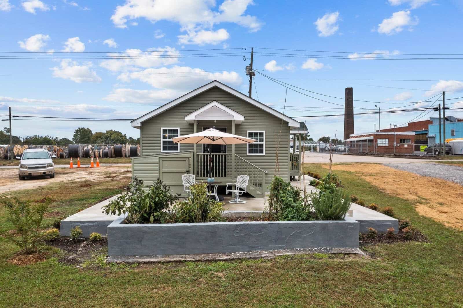 A small suburban house with a concrete front yard featuring a table and two chairs under an umbrella, flanked by minimalistic landscaping, with a vehicle parked to the side and industrial buildings in the background.