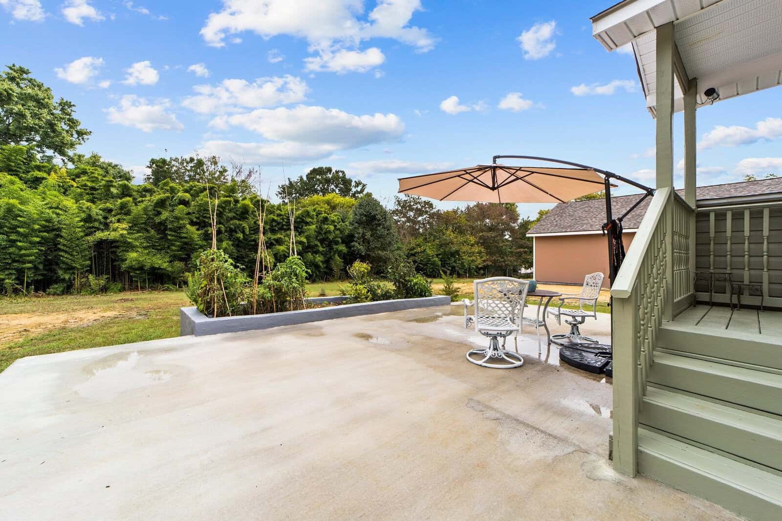 A residential patio with an umbrella-shaded table and chairs overlooking a green backyard and trees under a partly cloudy sky.