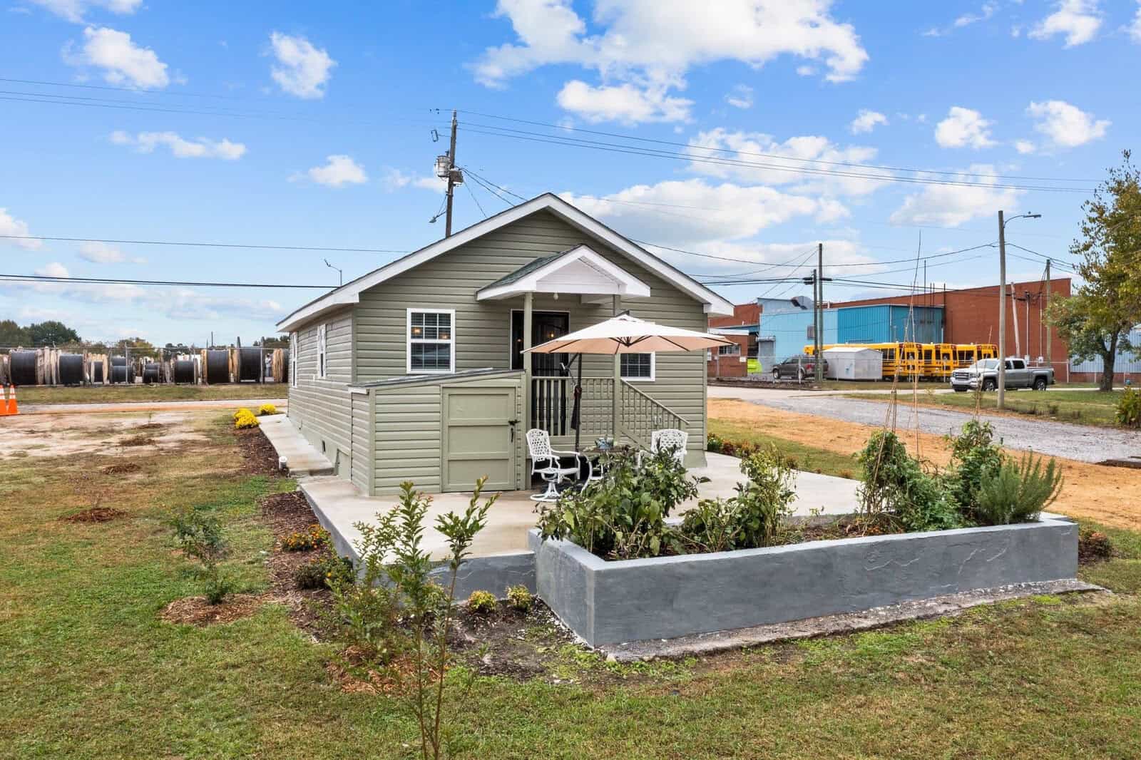 A small single-story house with beige siding is situated next to a raised garden bed with outdoor furniture, set against a backdrop of industrial train cars and a clear blue sky.