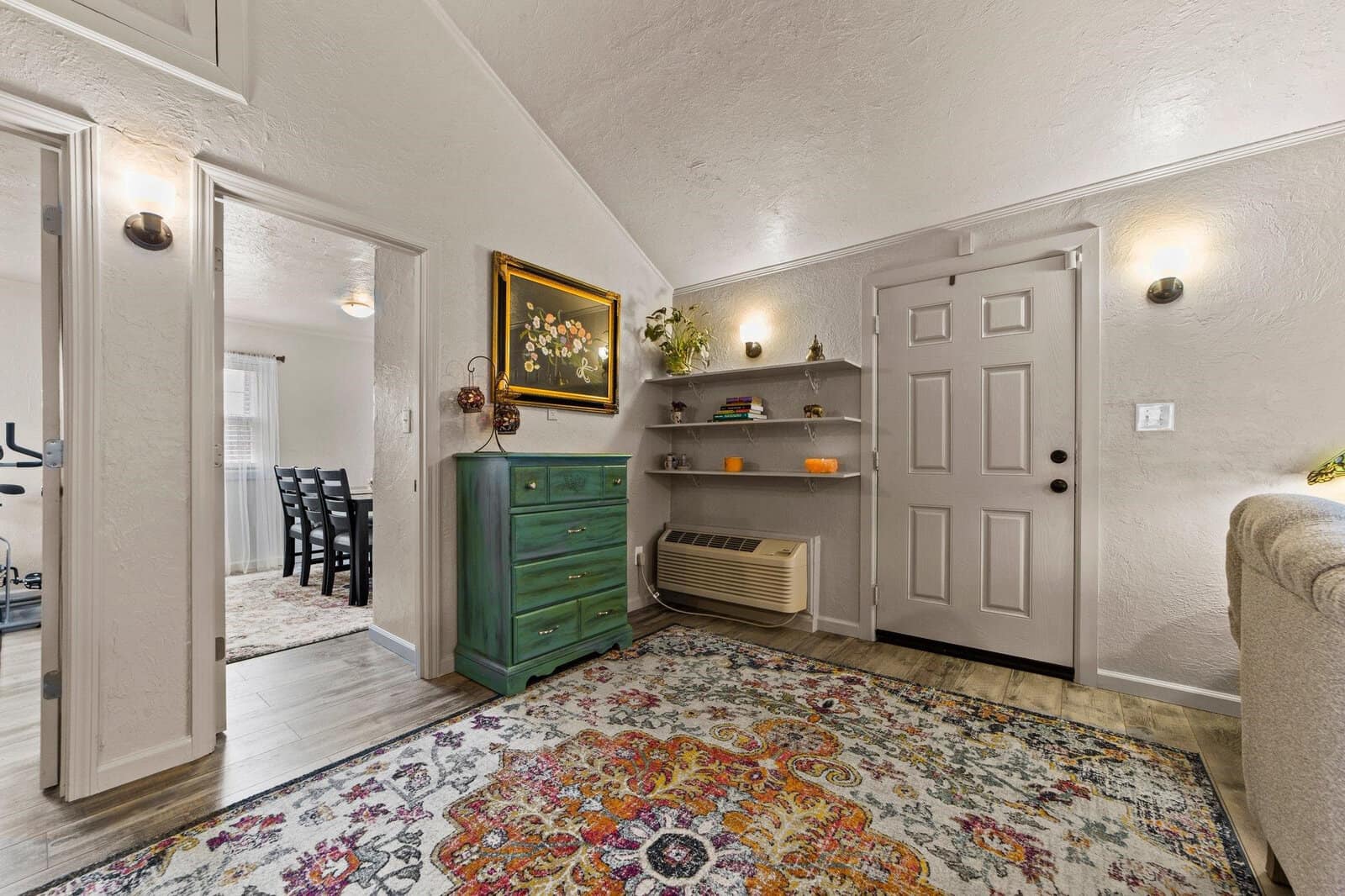 A warmly lit entryway featuring a patterned area rug, white walls, a green chest of drawers, floating shelves with decorative items, and a wall-mounted air conditioning unit, leading to an interior space with a dining table visible in the background.