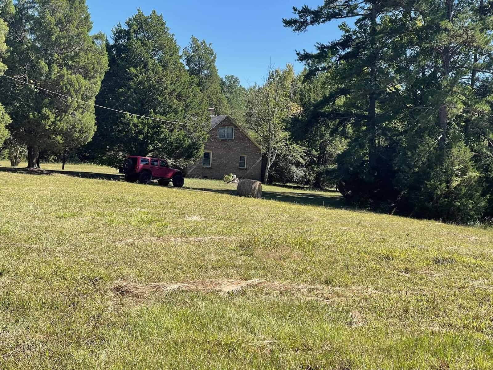 A red suv parked in the grass near a brick house with a hay bale in the foreground and tall trees in the background on a sunny day.