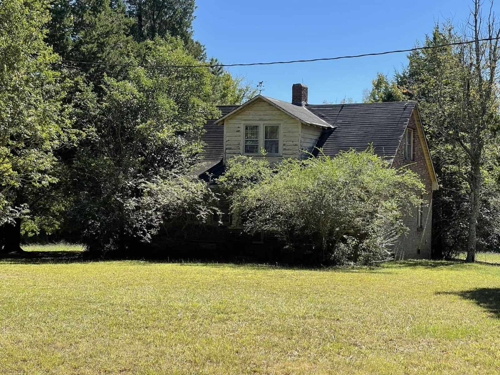 A weathered two-story house with overgrown bushes and trees in the front yard on a sunny day.