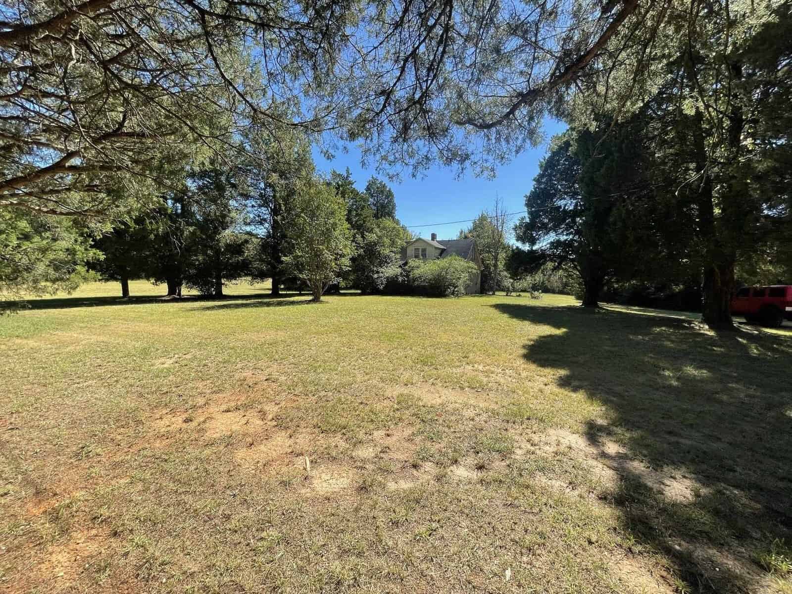 A tranquil rural landscape featuring a meadow with scattered trees, a house in the background, and a red vehicle partially visible to the right, under a clear sky.
