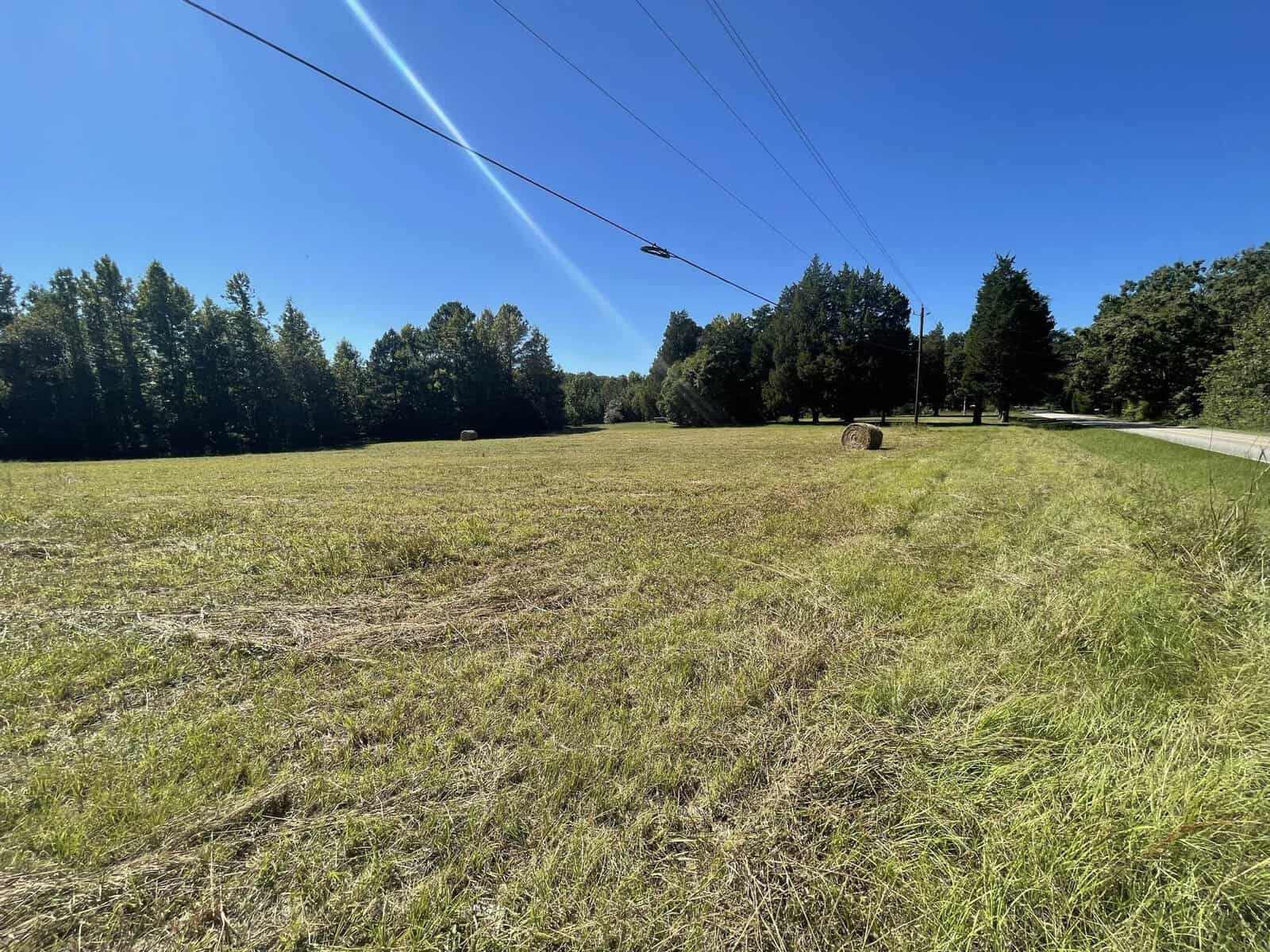 A clear, sunny day over a mowed field with a power line stretching across, a road visible on the right, and a backdrop of dense trees.