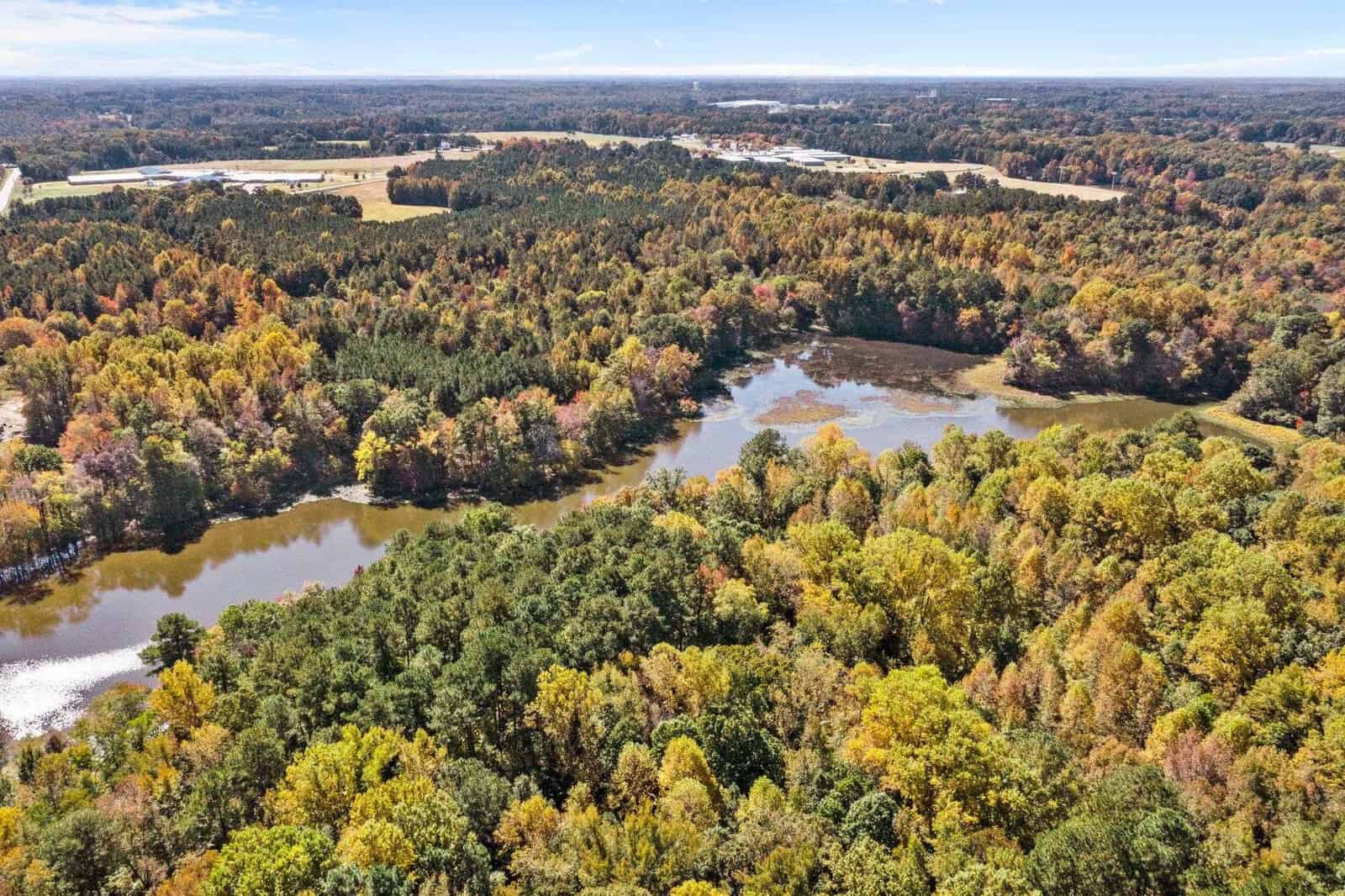 Aerial view of a landscape featuring a mosaic of autumn-colored trees surrounding a meandering body of water, with distant buildings indicating the proximity of a developed area.
